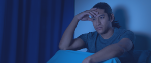 A man sitting up, awake in bed illuminated by dim blue light.