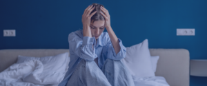 A young woman crouched on her bed with her hands on her head.